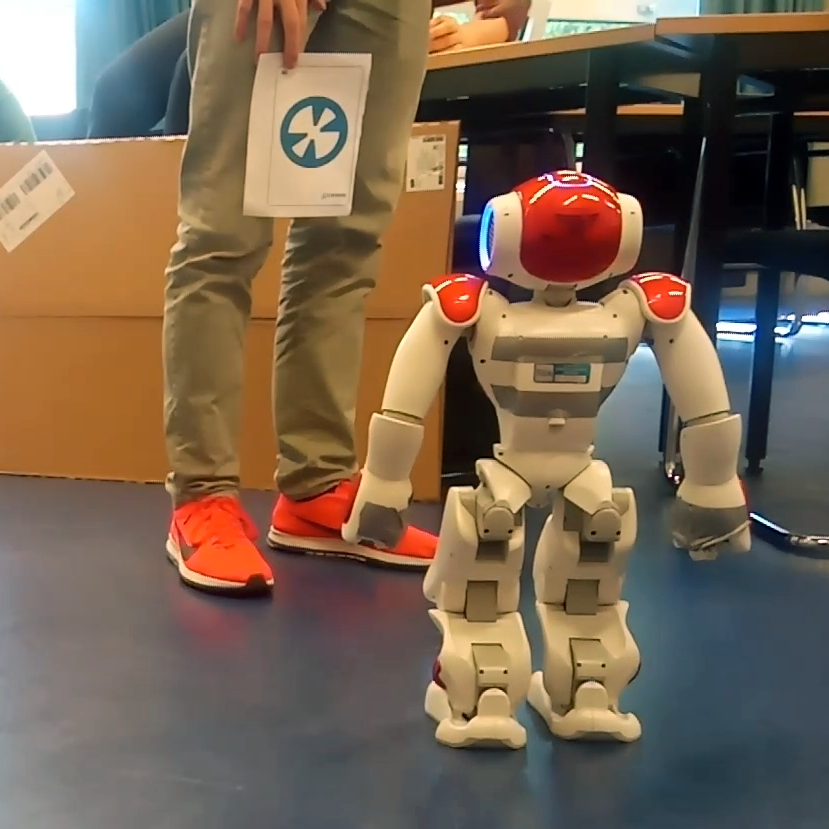 The NAO robot seen from the back. A person stands in front of it, holding a piece of paper with a landmark for it to scan. 
